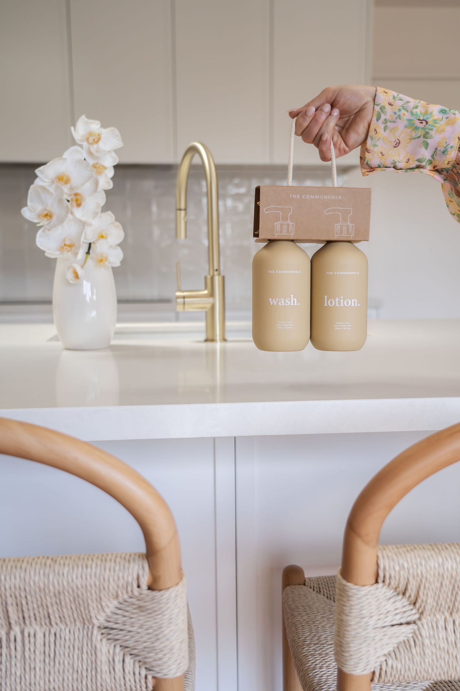 Modern kitchen counter with gold faucet, white orchids, and two beige bottles beside rattan bar stools.