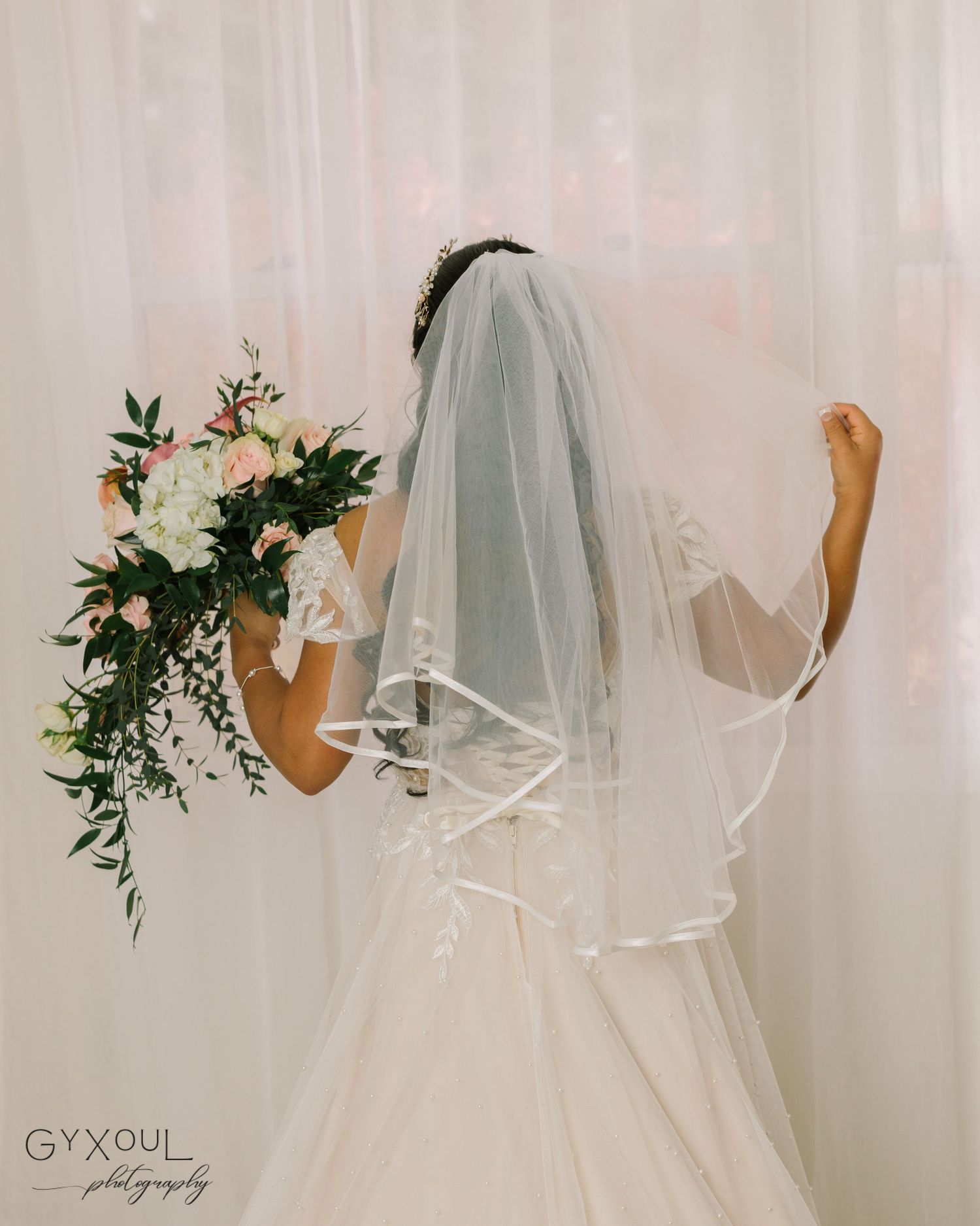 Back view of bride in white dress holding bouquet against light backdrop.