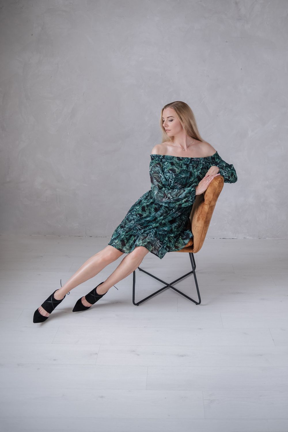 Elegant pose in green floral dress on designer chair against gray background.