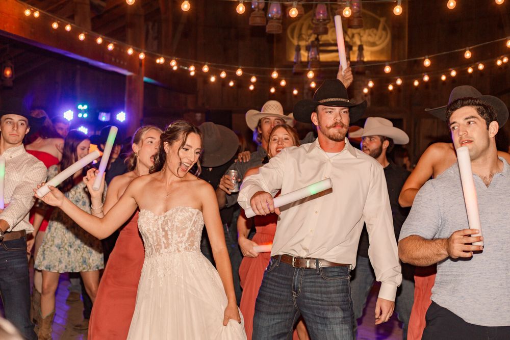 Wedding guests dancing in a line formation at a barn reception with string lights overhead.