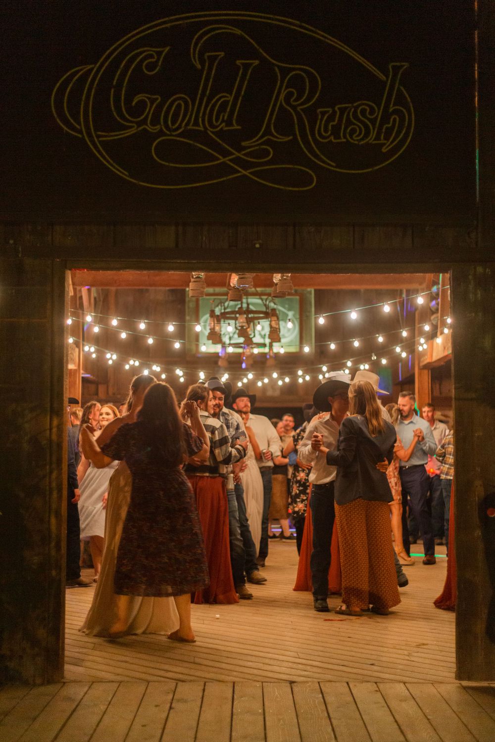 Couples dancing at a rustic venue with 'Gold Rush' signage and string lights overhead.