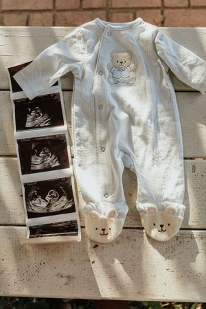 Baby announcement featuring a light blue teddy bear onesie alongside ultrasound photos laid on a white wooden surface.