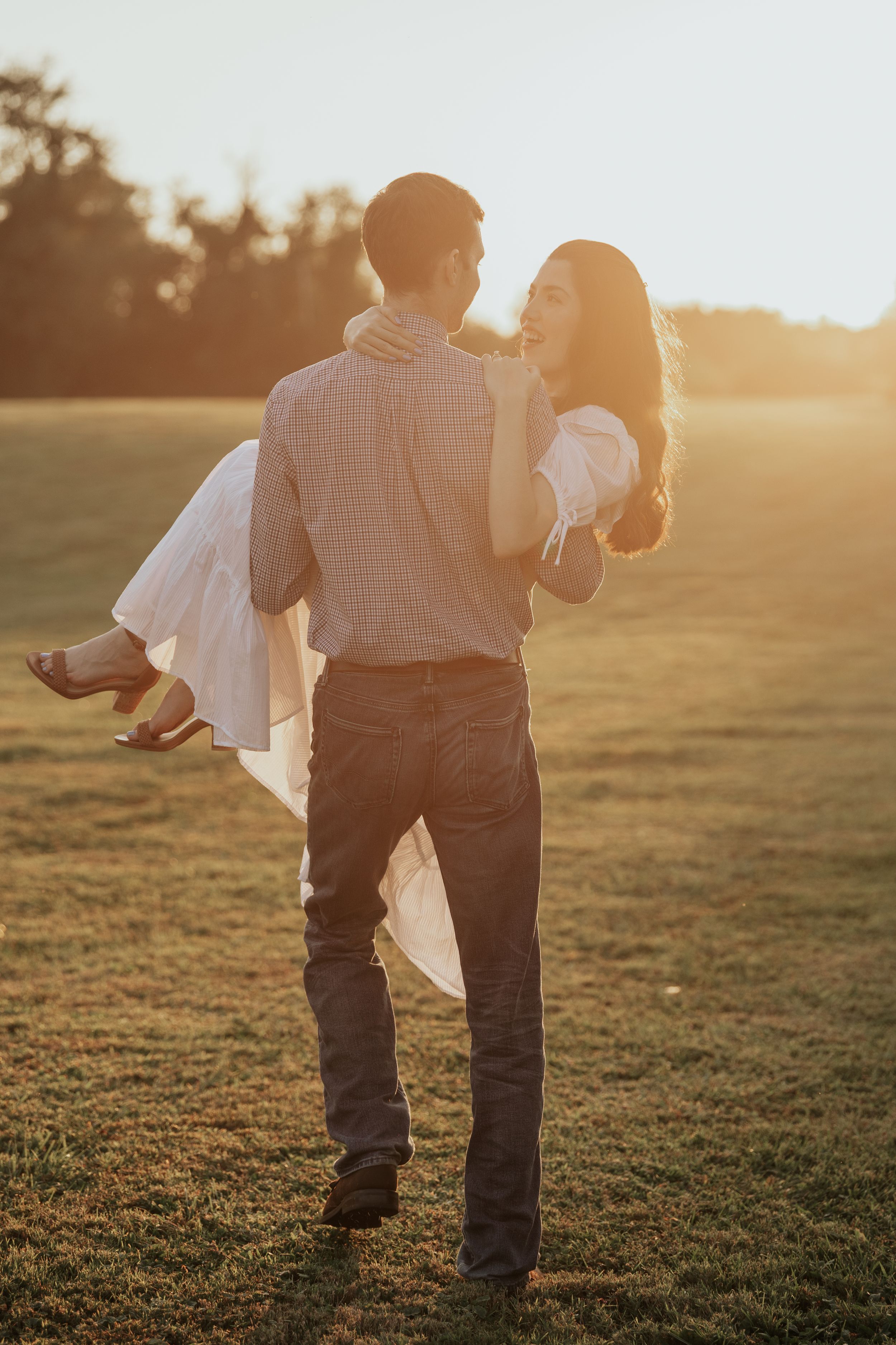 A couple embraces in a sunlit field during golden hour, with the person in white being carried romantically at sunset.