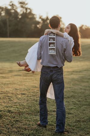 A couple stands together in a grassy field at sunset holding an ultrasound photo to announce their pregnancy.