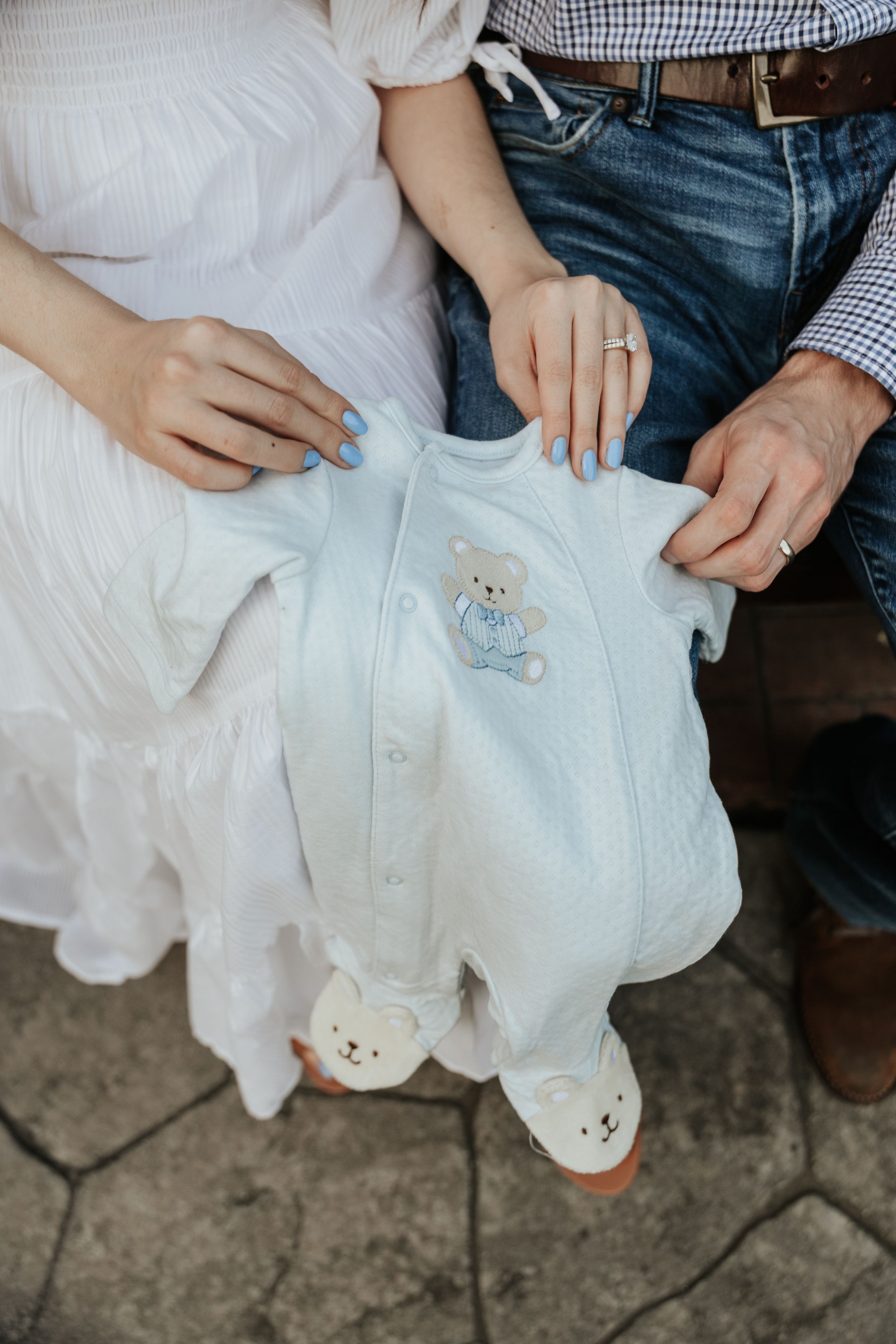 Hands holding a light blue baby onesie with teddy bear embroidery and footie details.