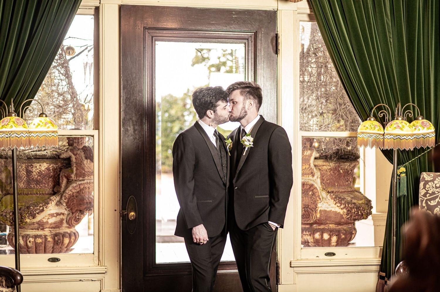 Two grooms in black suits sharing a kiss at their wedding ceremony in an elegant decorated venue.