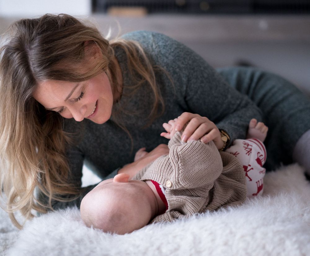 Ein Elternteil in einem grauen Pullover teilt einen zärtlichen Moment mit seinem Baby, das auf einer weißen, flauschigen Decke liegt.