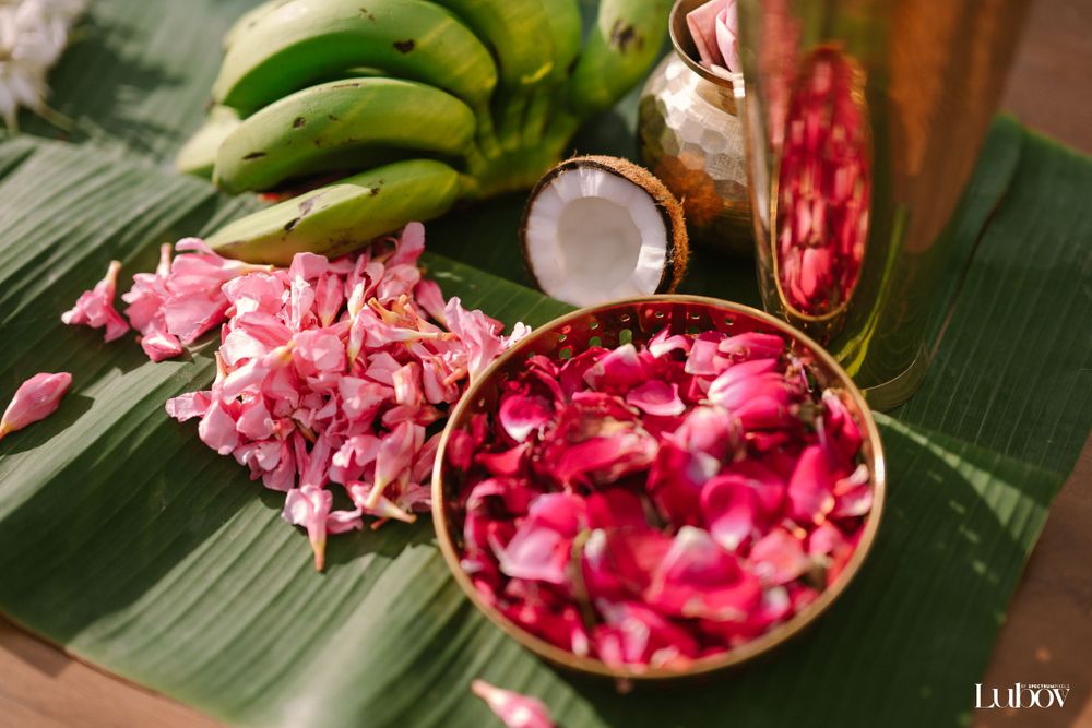 Pink rose petals and coconut arranged on a banana leaf with tropical fruits in the background.