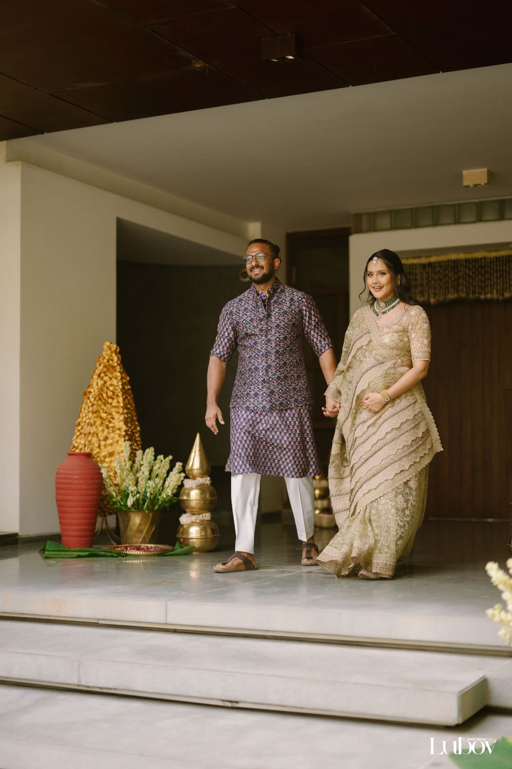 A couple in traditional Indian attire stands on a porch with decorative elements including a golden tree and pottery.