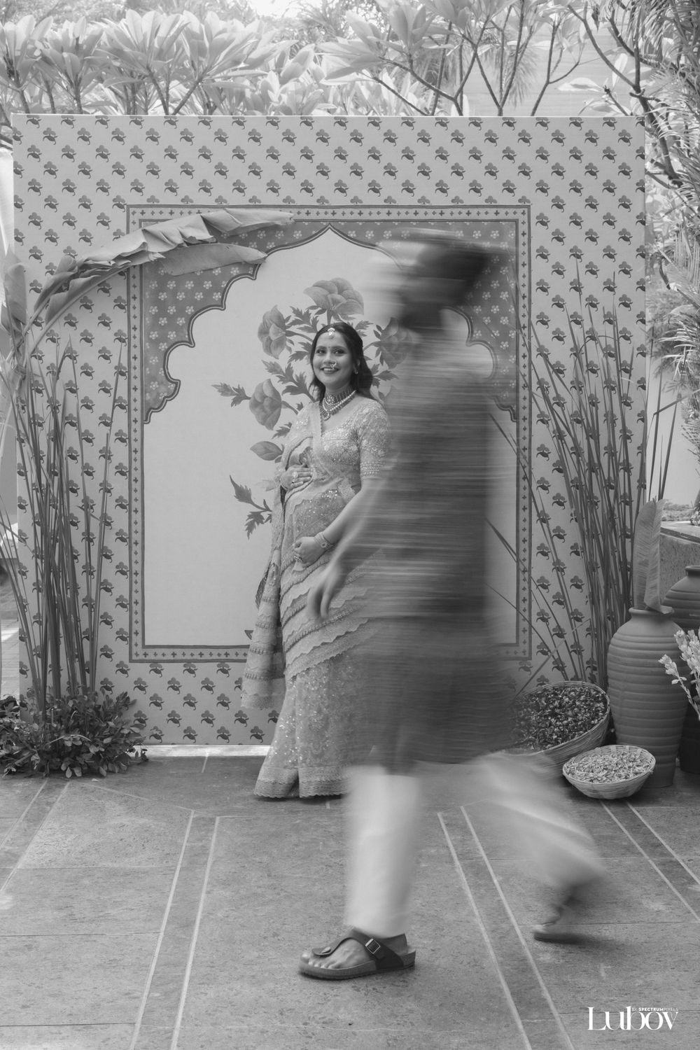 A person in a cream saree poses against a patterned backdrop with bamboo decor.