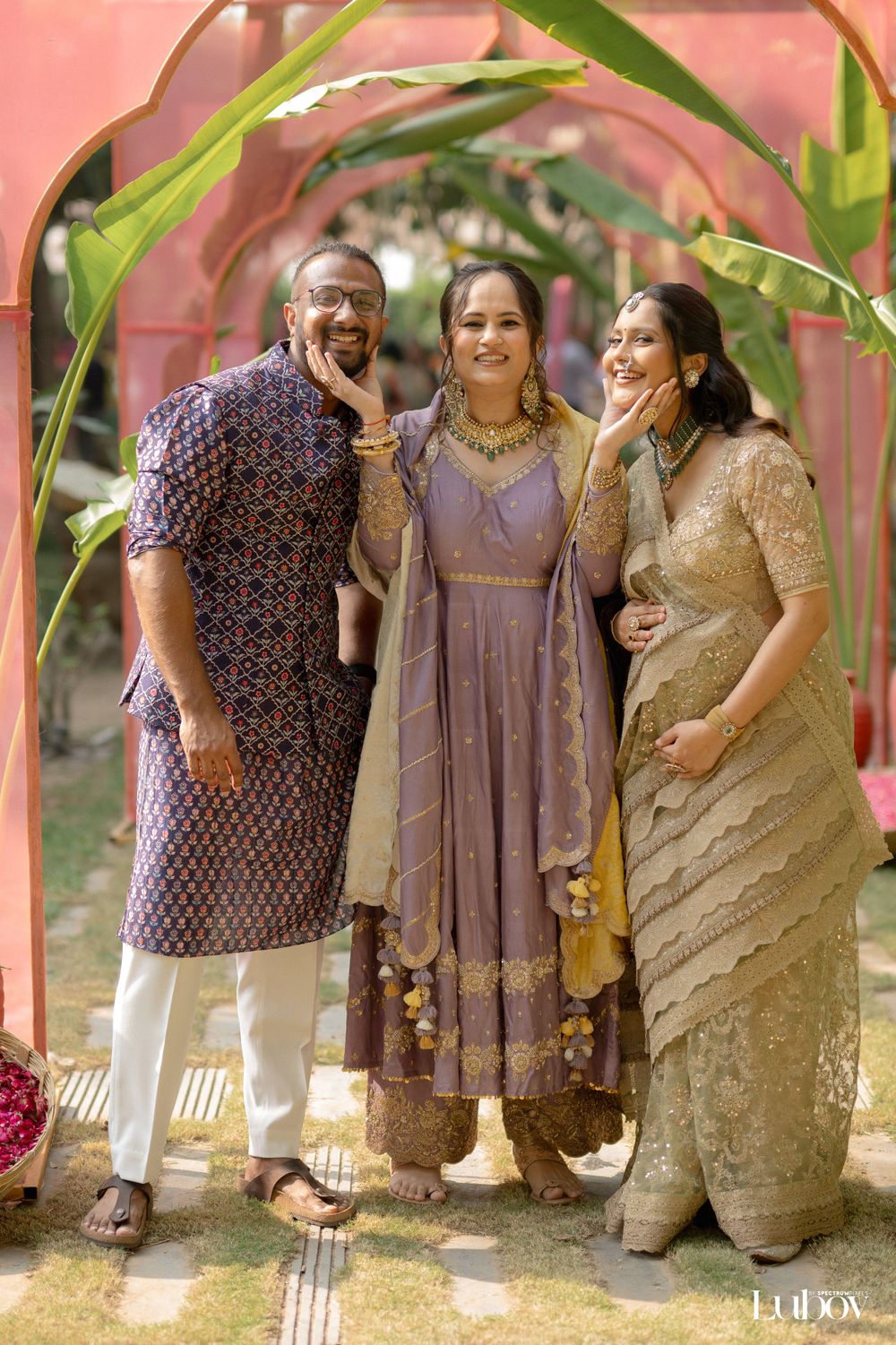 Three people in traditional Indian outfits pose together in a garden setting with pink walls.