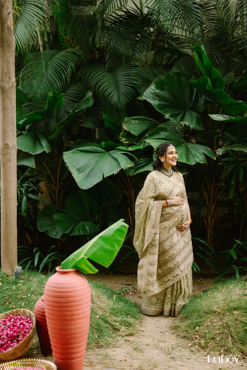 A person in a beige saree poses elegantly against lush tropical foliage and a terracotta pot.