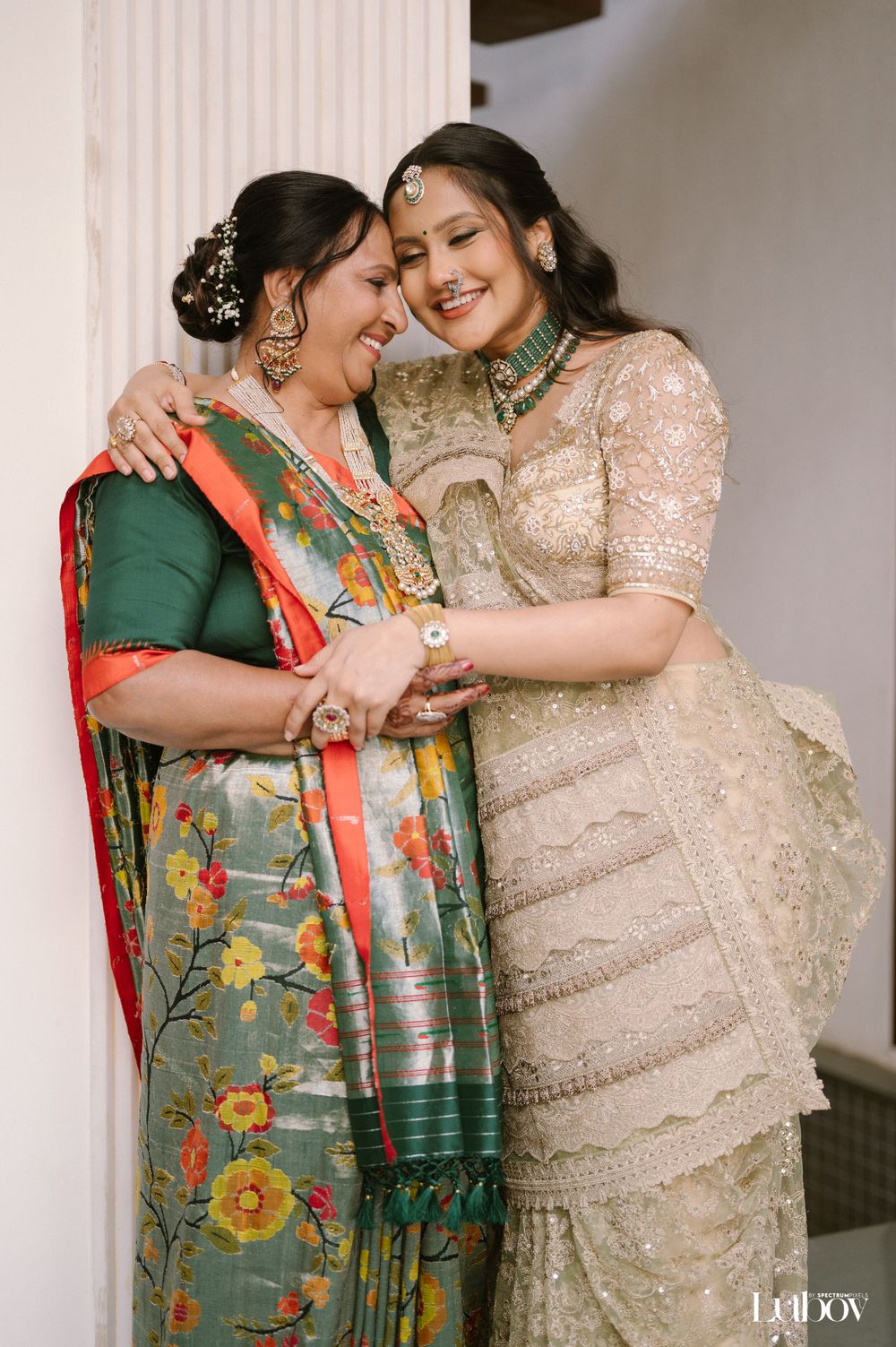 Two people in traditional Indian sarees share a joyful embrace against a white wall.