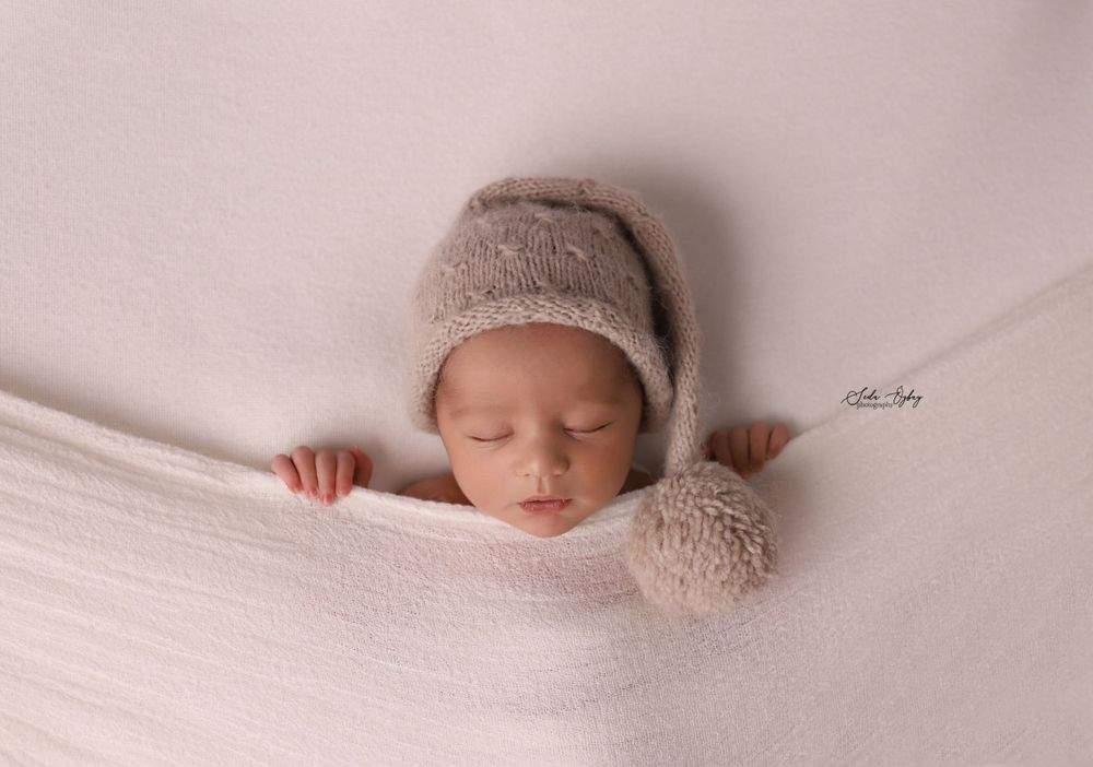 Peaceful newborn wrapped in soft white fabric with knit bonnet against pink background.