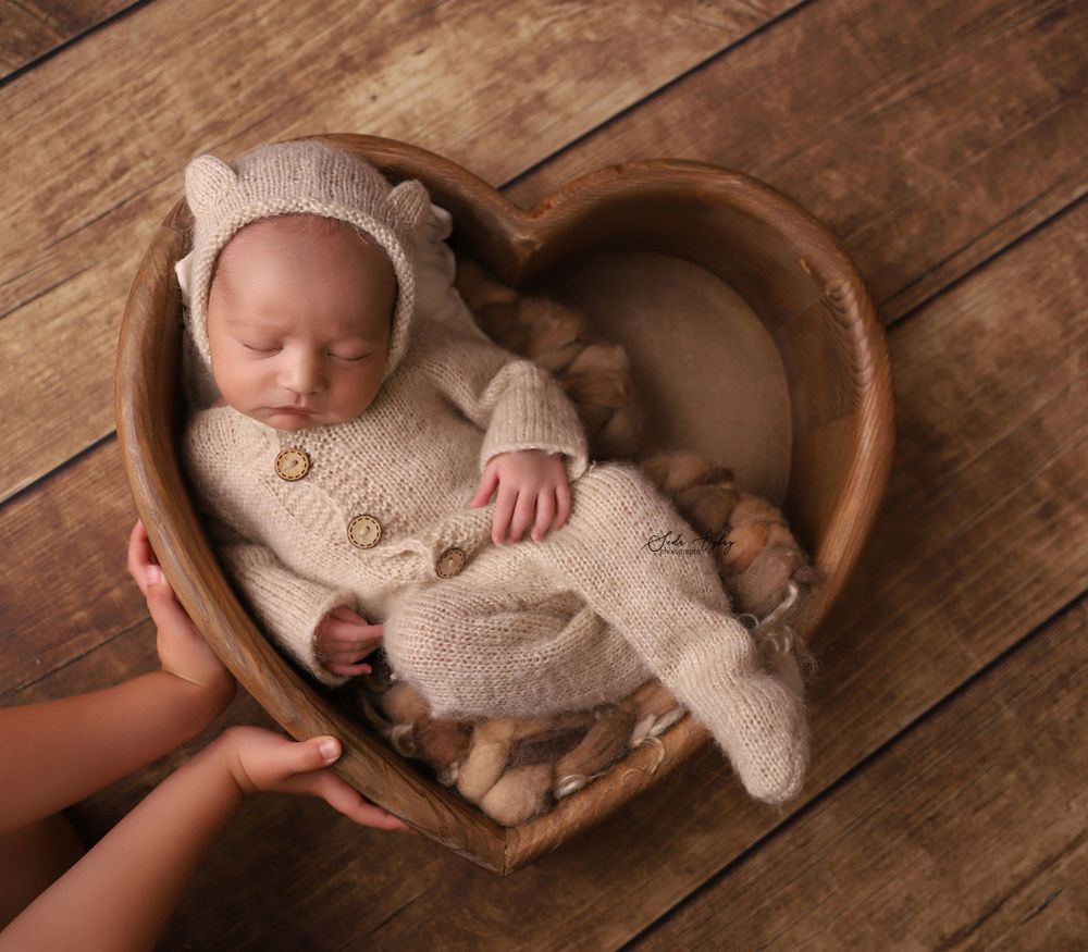 Sleeping newborn in cozy knit outfit nestled in heart-shaped wooden bowl on rustic wooden floor.