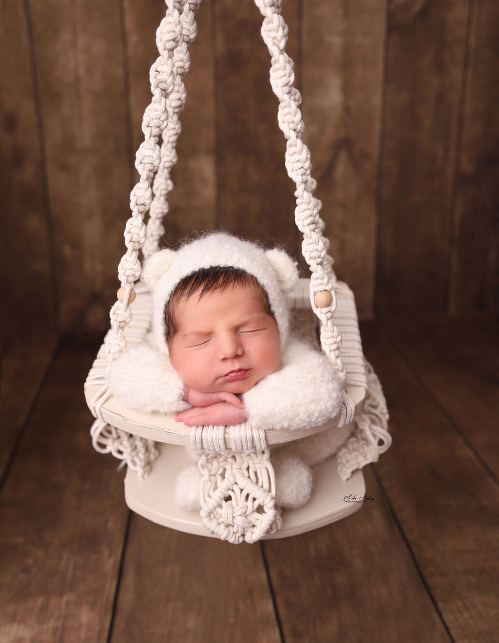 Sleeping newborn in white macrame hanging basket against wooden backdrop.