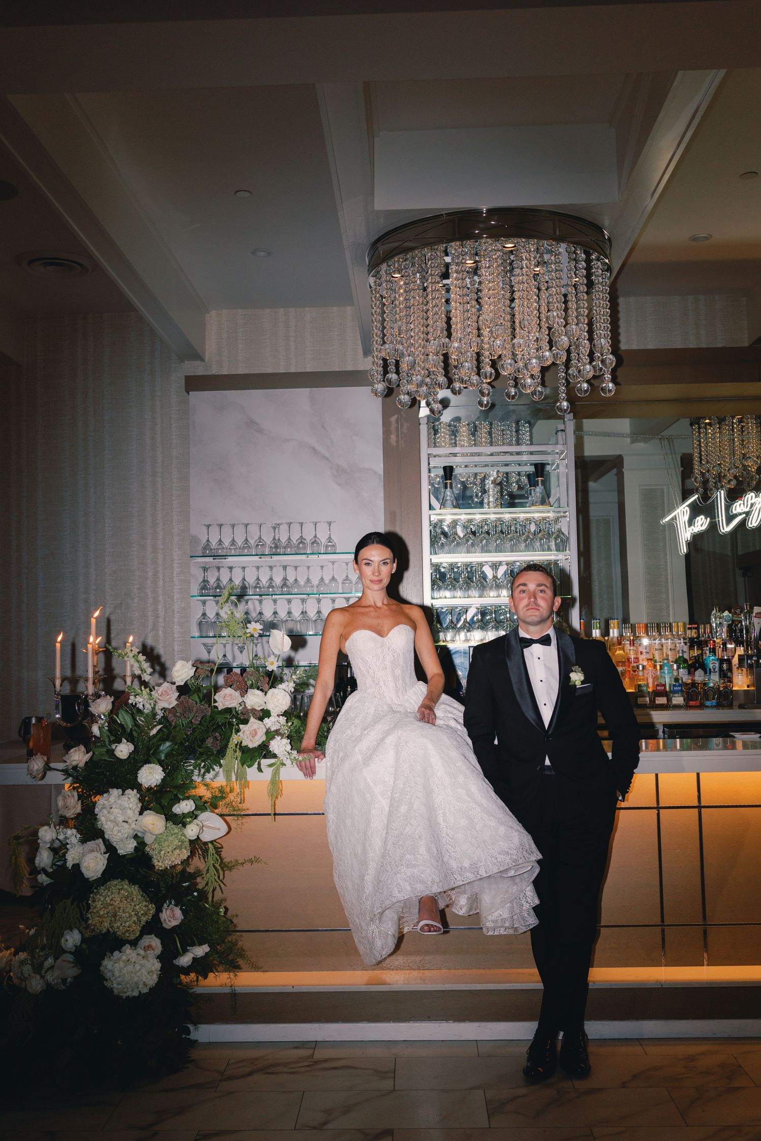 Bride in strapless gown and groom in tuxedo posing at an elegant bar with crystal chandelier above.