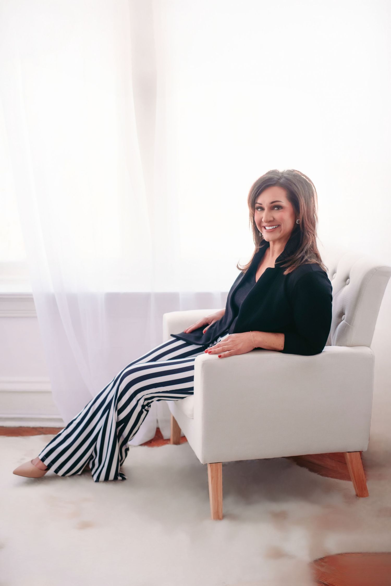 Sitting in white armchair wearing black blazer and striped pants with neutral decor and soft lighting in background.