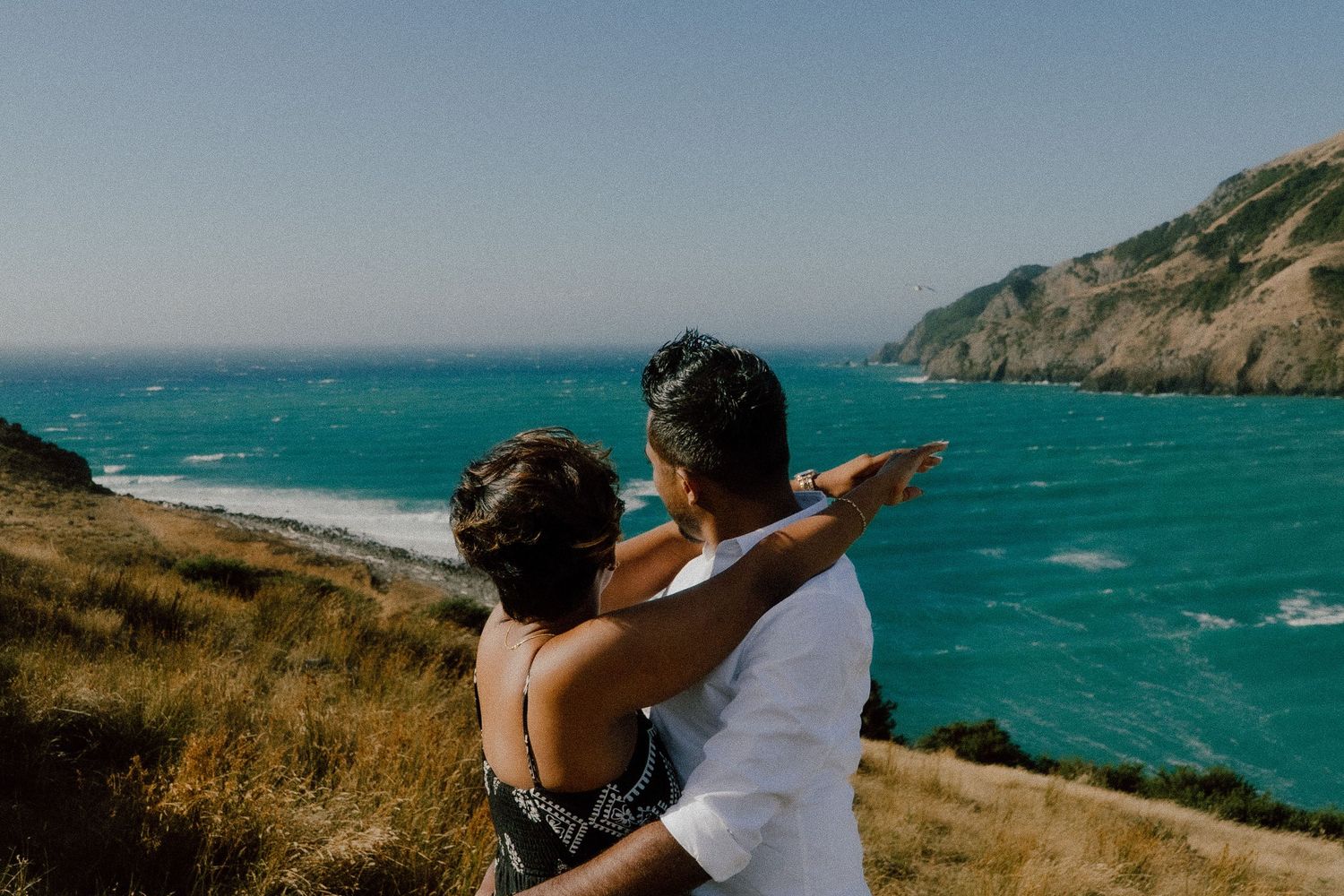 Couple embracing on a scenic coastal overlook, pointing toward turquoise ocean waters with green hills in the background.