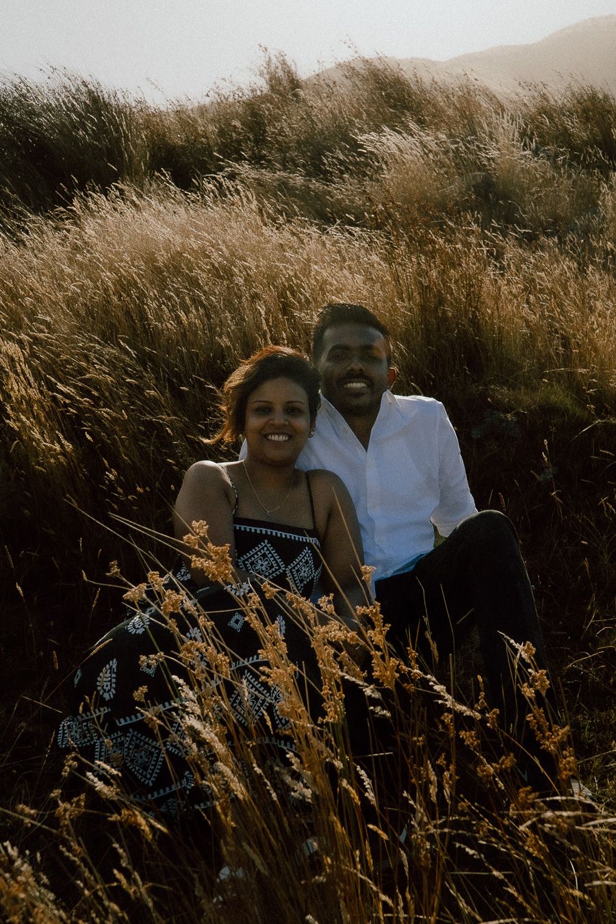 A smiling couple sits together in golden tall grass fields, enjoying a romantic outdoor photo session.