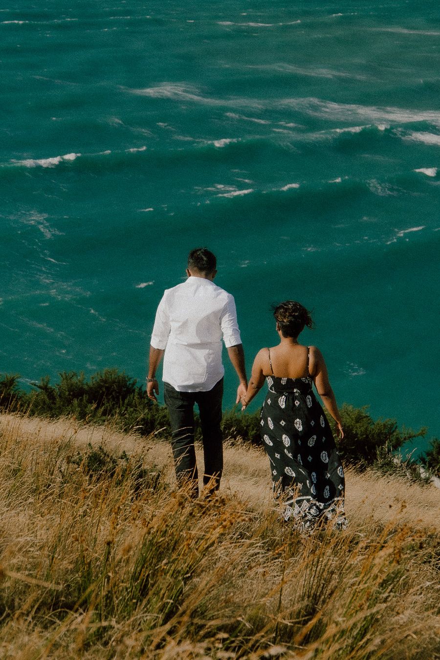 Couple holding hands walking along a coastal cliff path overlooking turquoise ocean waves.