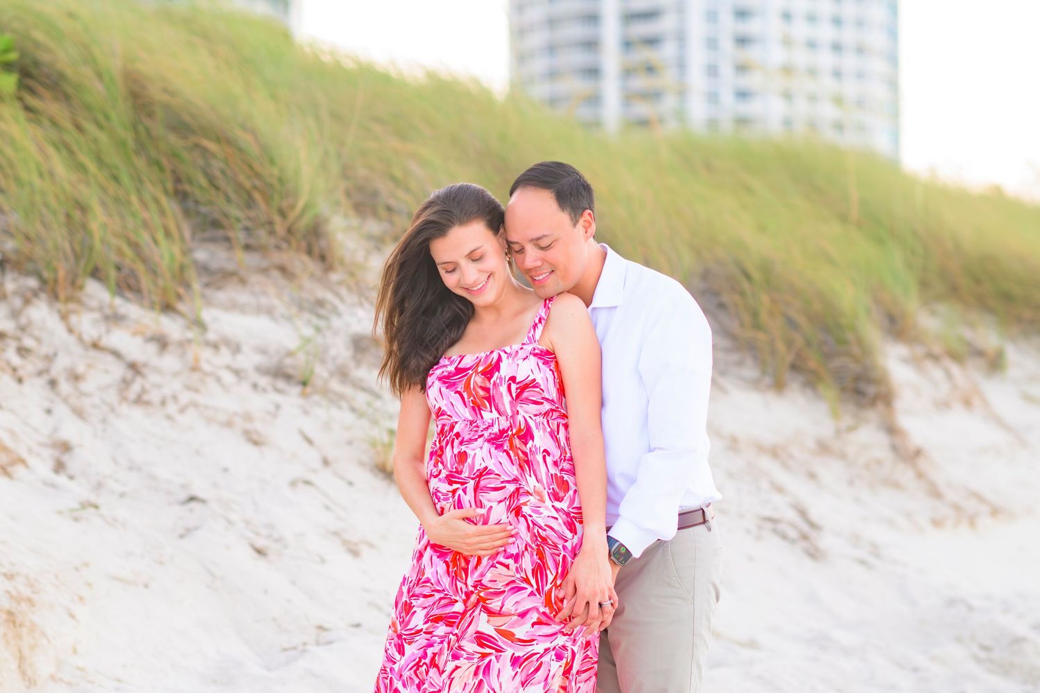 A couple in love embrace on a beach with sand dunes and a coastal high-rise building in the background.