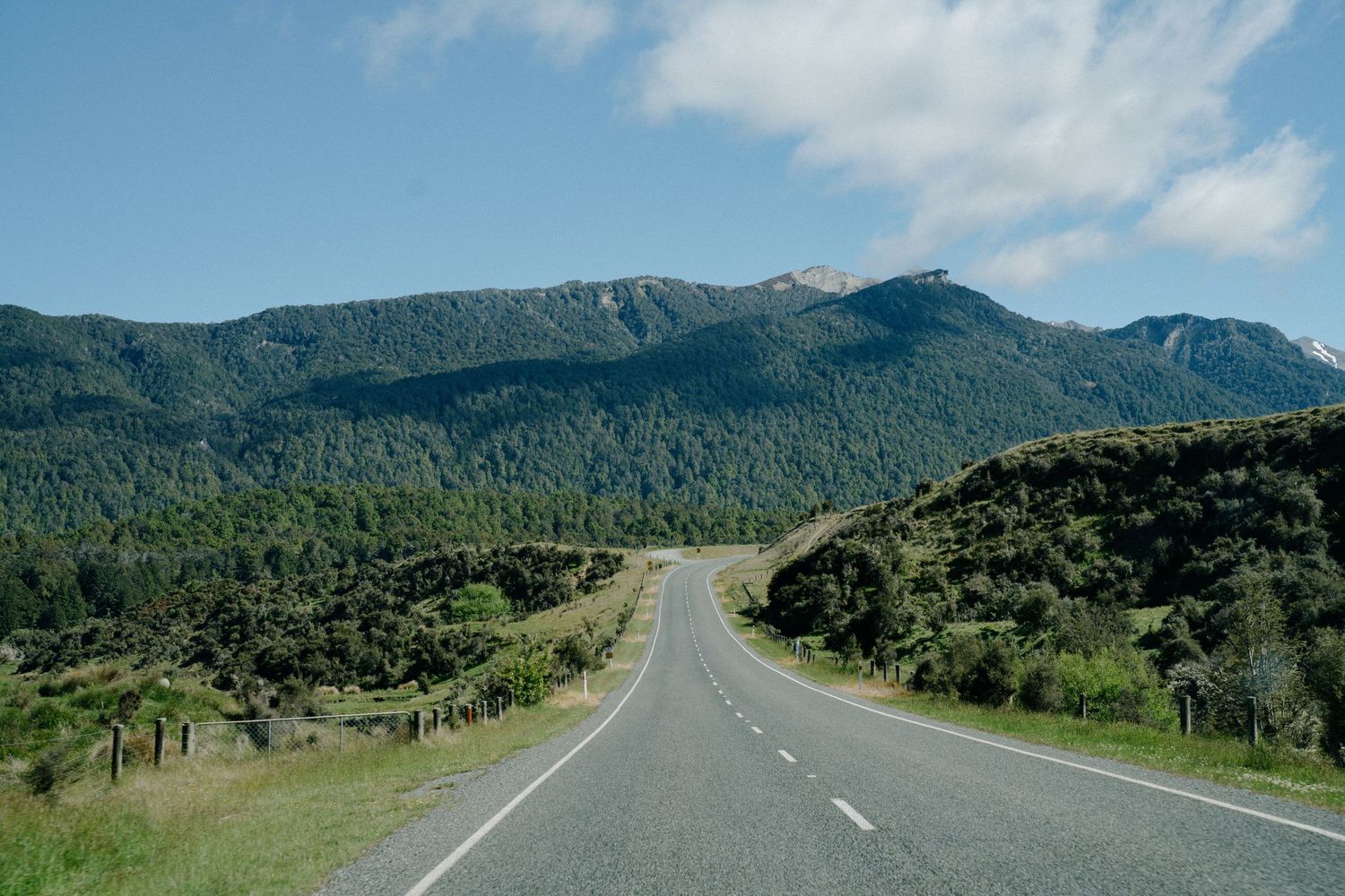 A scenic mountain road winding through green hills and forest under a blue sky.