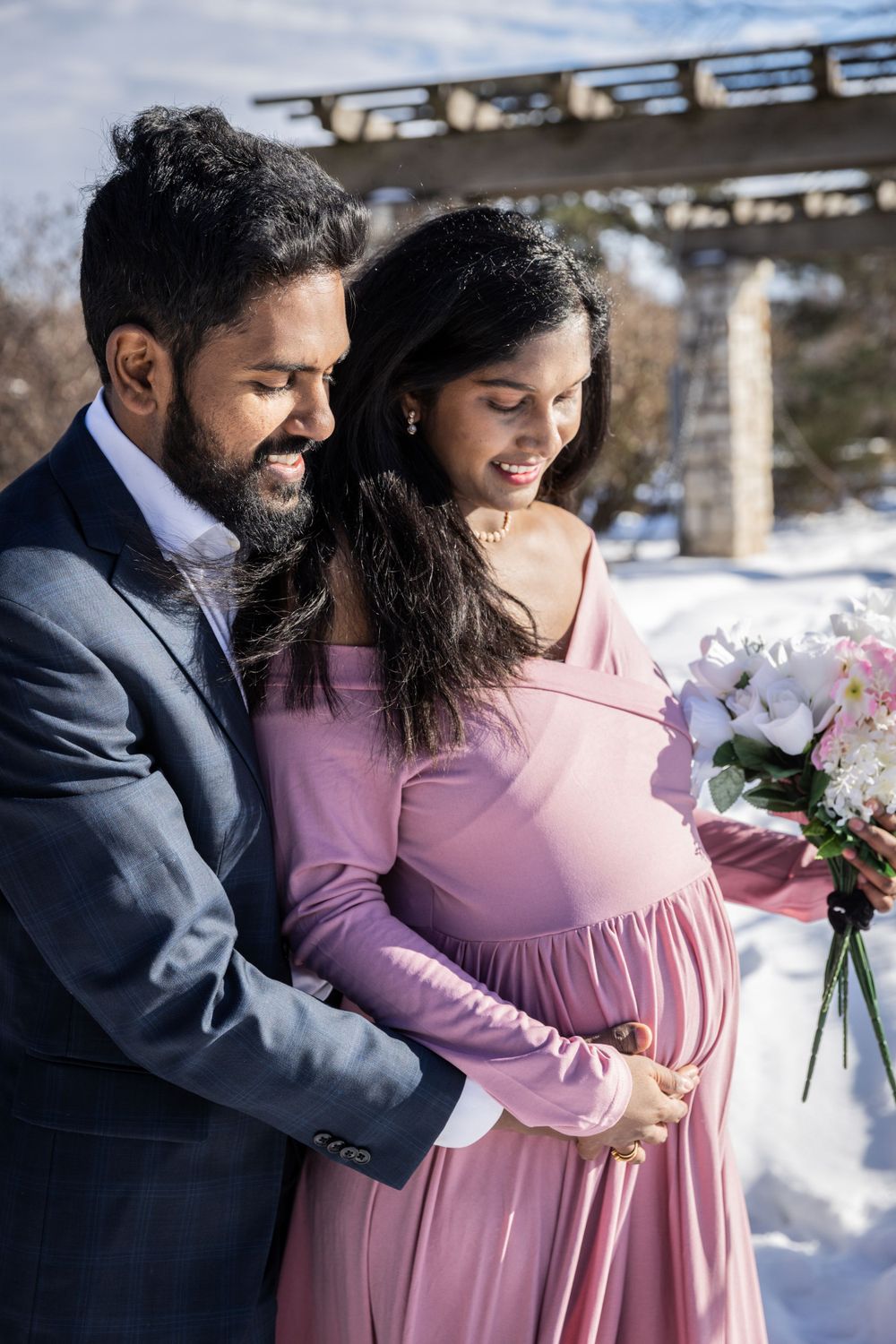 Couple shares intimate moment in pink dress and navy suit against snowy winter backdrop.