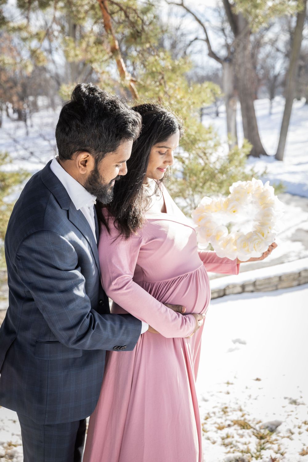 A couple shares an intimate winter maternity photoshoot moment in the snow with the woman wearing a pink dress.