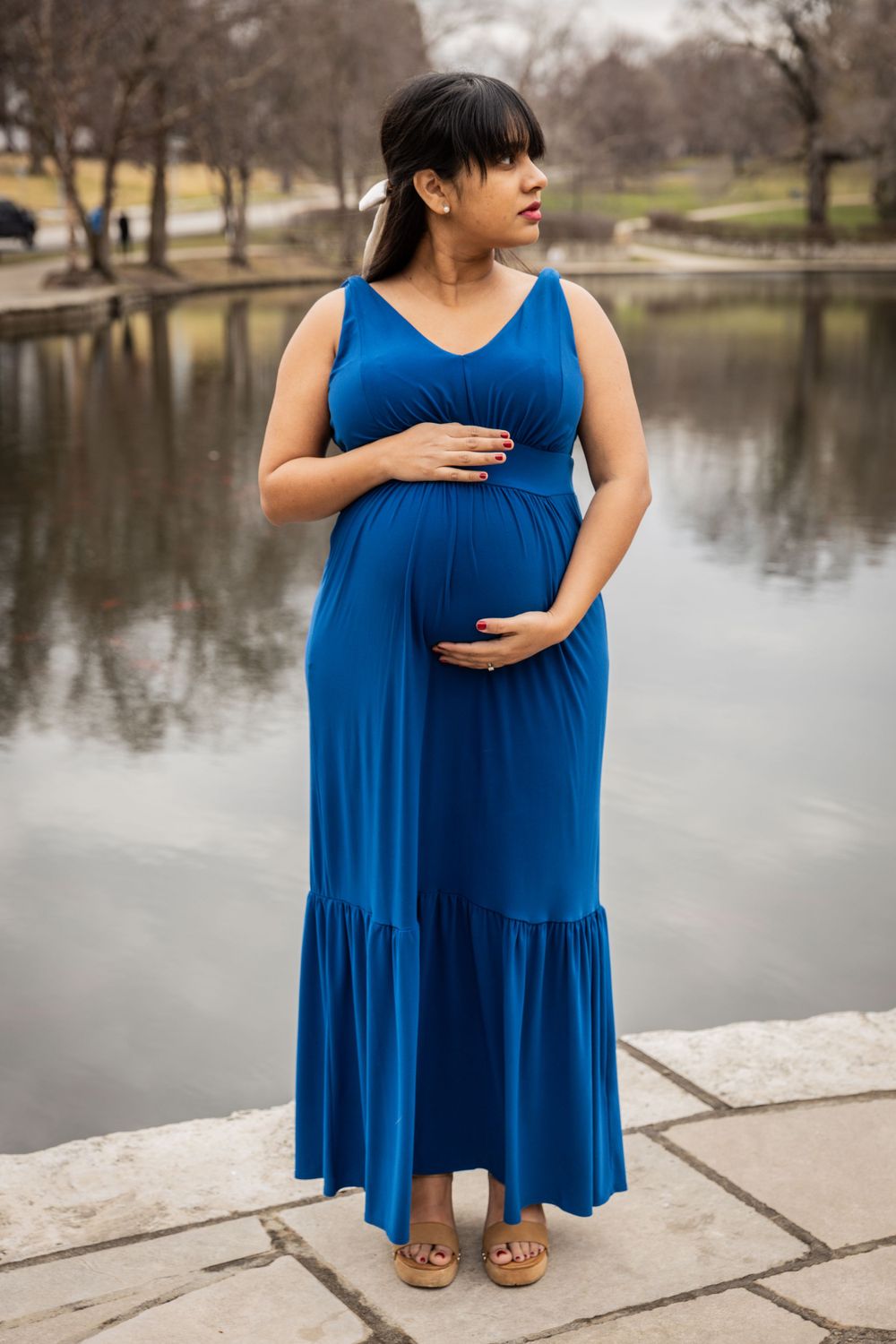 Woman in flowing blue dress stands by lakeside during maternity photoshoot.
