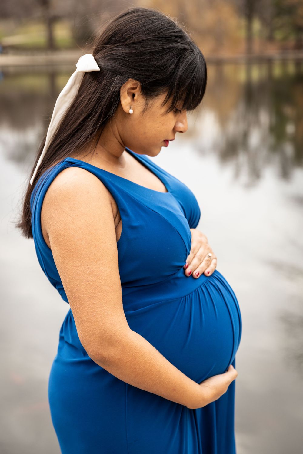 Woman in blue dress stands by lake during maternity photoshoot.