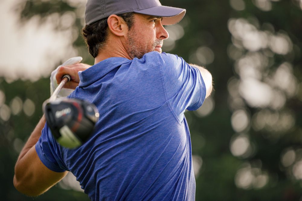 Golfer mid-swing in blue shirt during afternoon light.