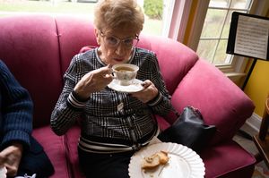 Elderly woman sips tea from fine china cup while seated in pink armchair at gathering.
