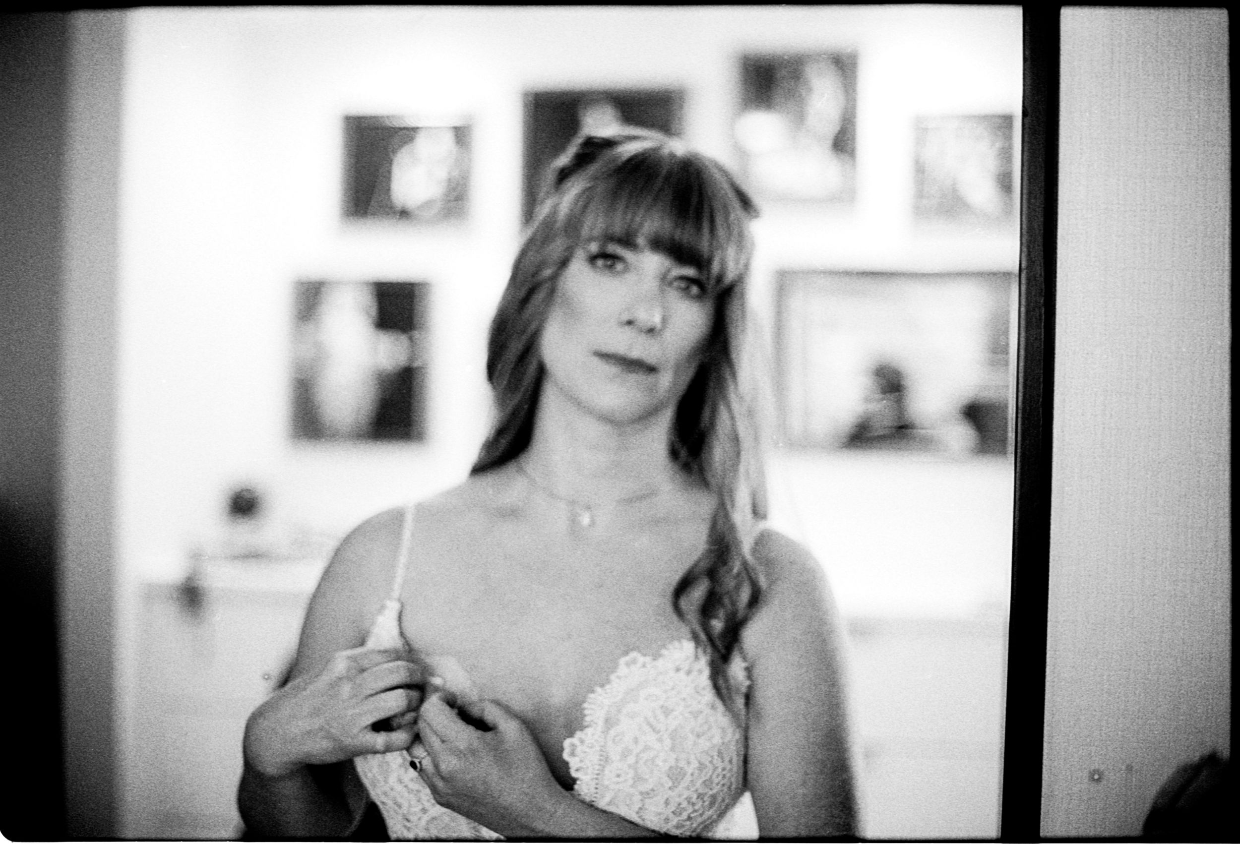 Black and white portrait of a woman in a lacy bra adjusting her outfit with framed photos on the wall behind her.