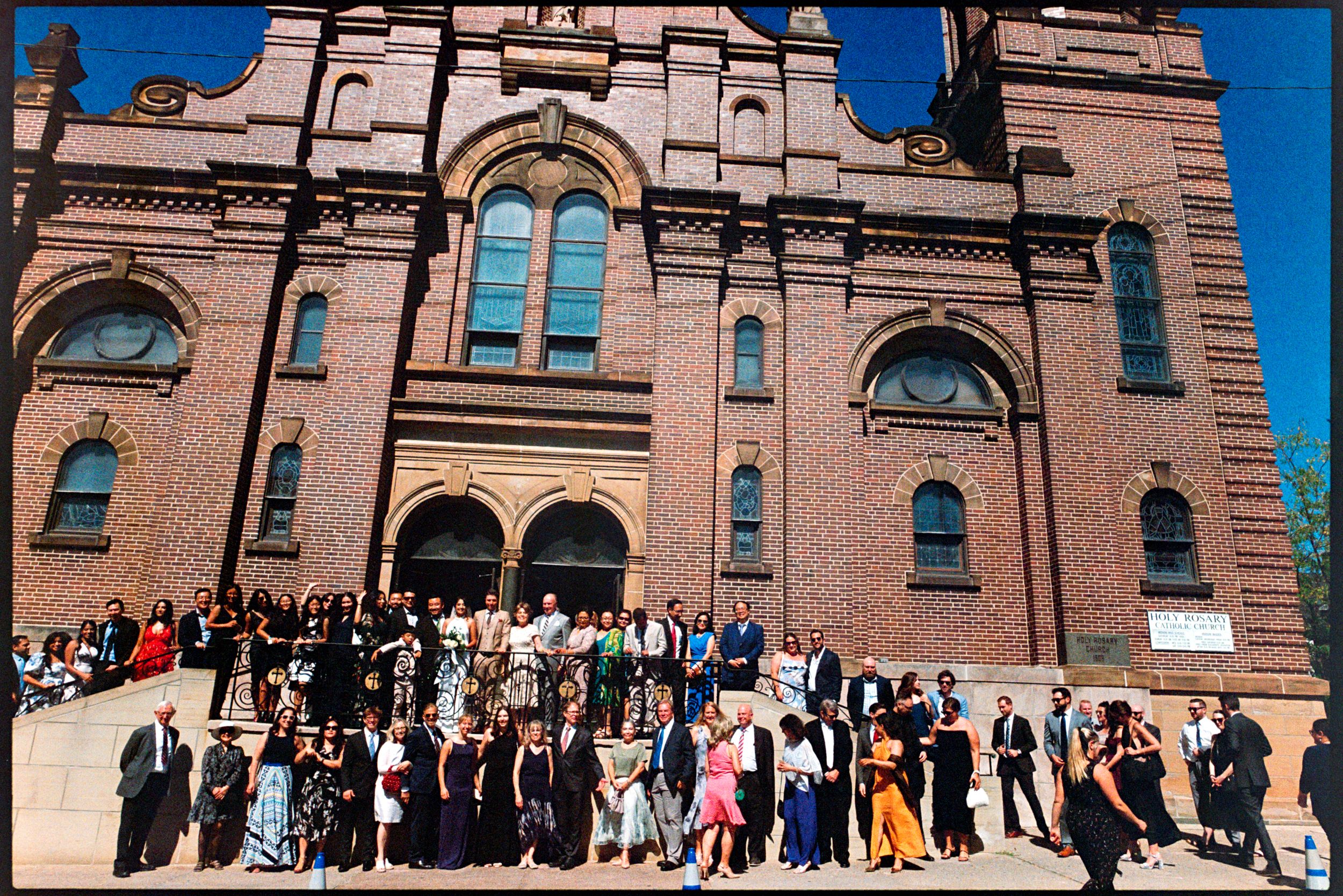 Large wedding party poses on steps of grand brick church with arched windows.