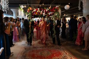Indian wedding ceremony with bride in pink saree walking under floral mandap arch amid misty aisle decor.