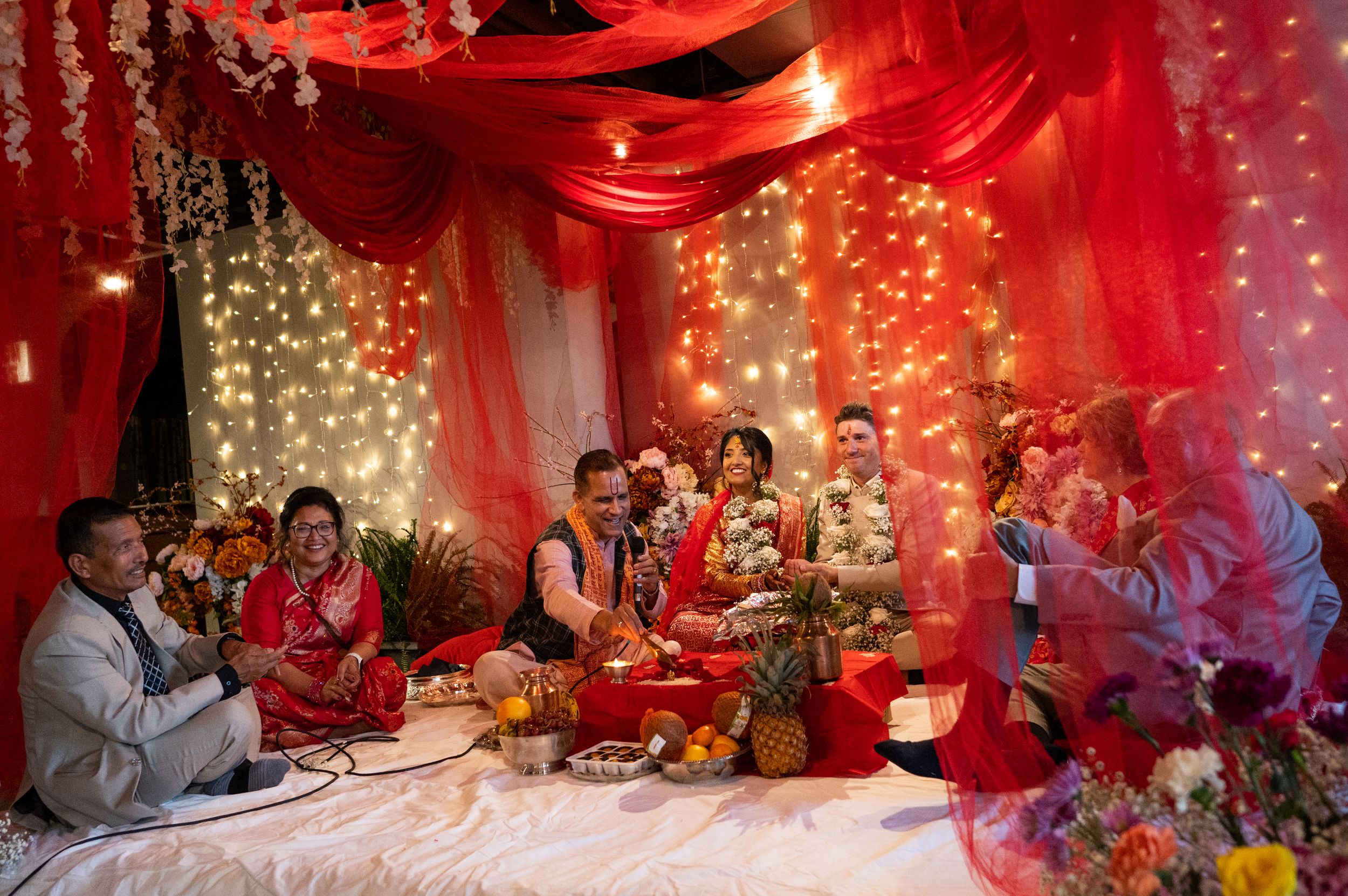 Traditional Indian wedding ceremony with bride and groom seated under red draped mandap with fairy lights.