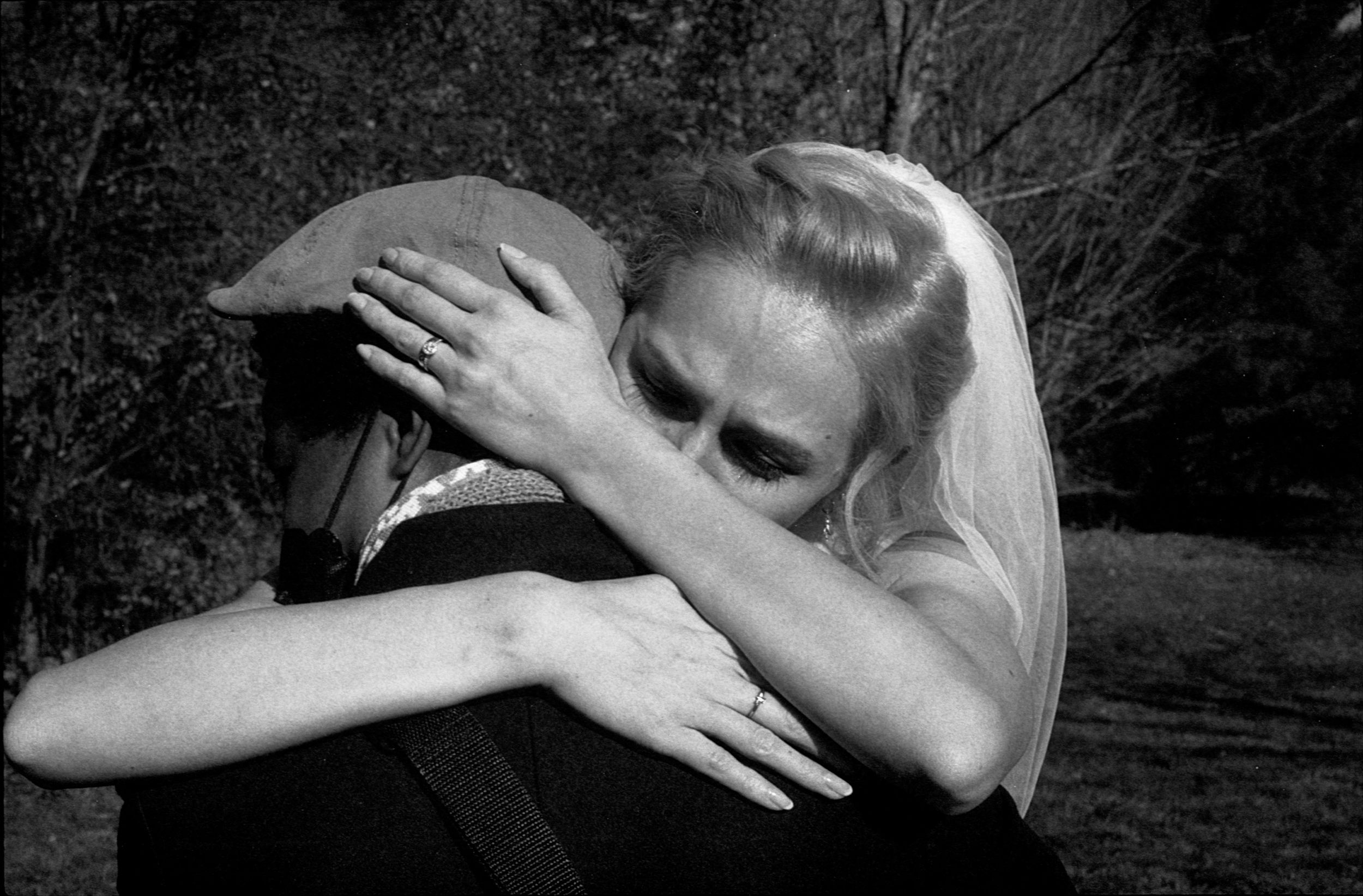 Black and white photo of a bride in a veil sharing an emotional hug outdoors among trees.