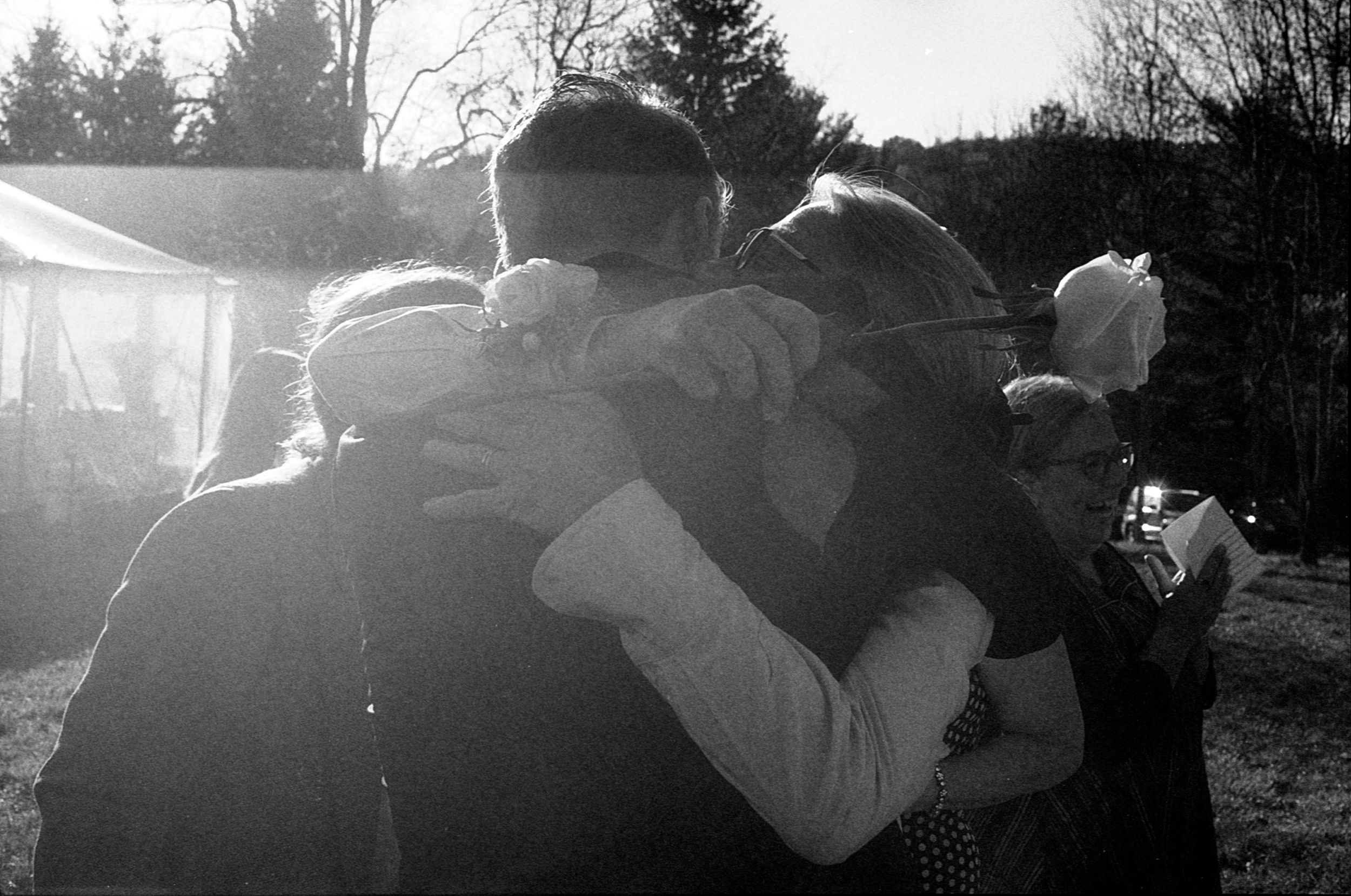 Black and white photo of a group of people sharing a warm embrace outdoors in soft sunlight.