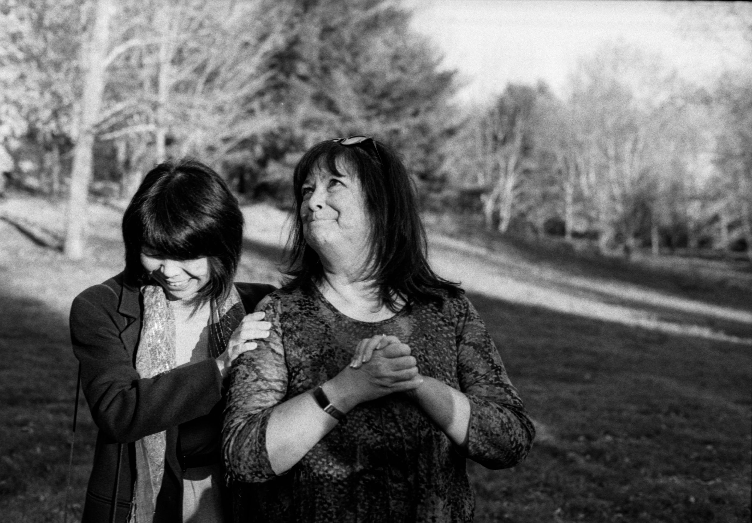 Two women sharing emotional moment outdoors among bare trees in black and white photo.