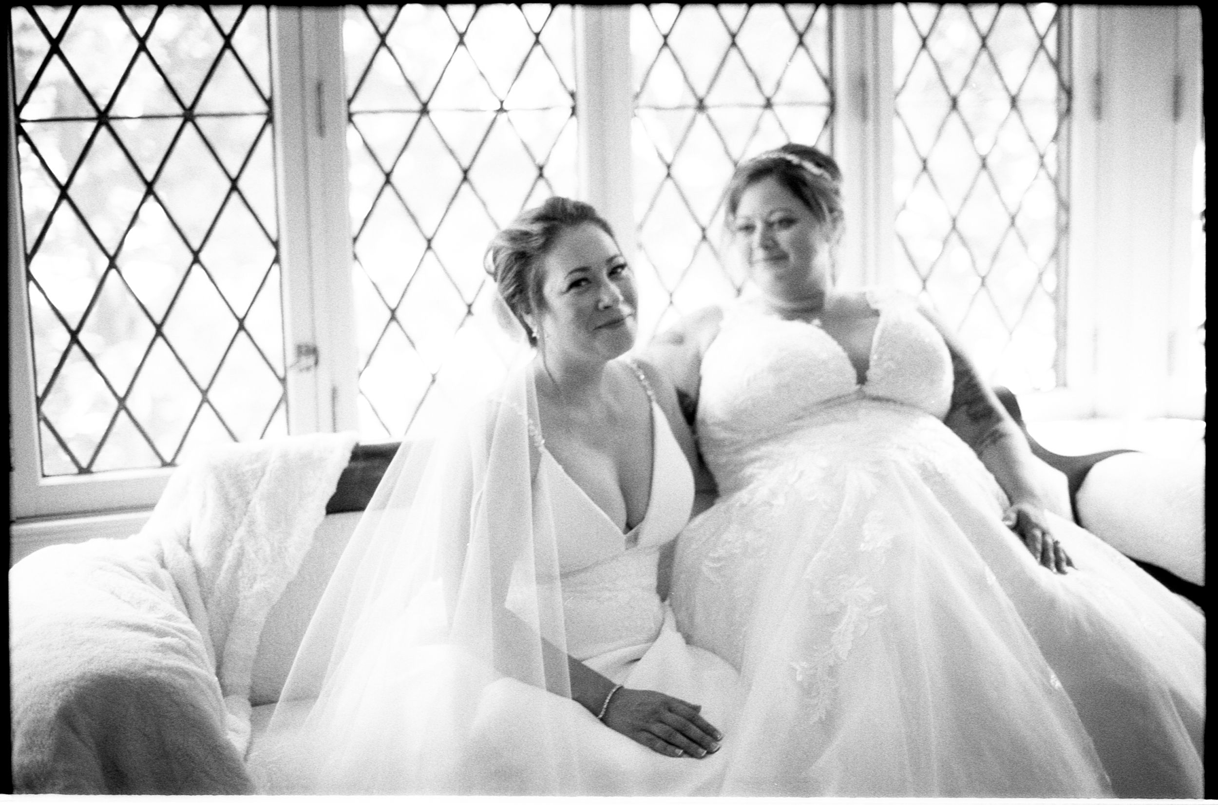 Two brides in white wedding gowns sit together near leaded glass windows in a black and white wedding photo.