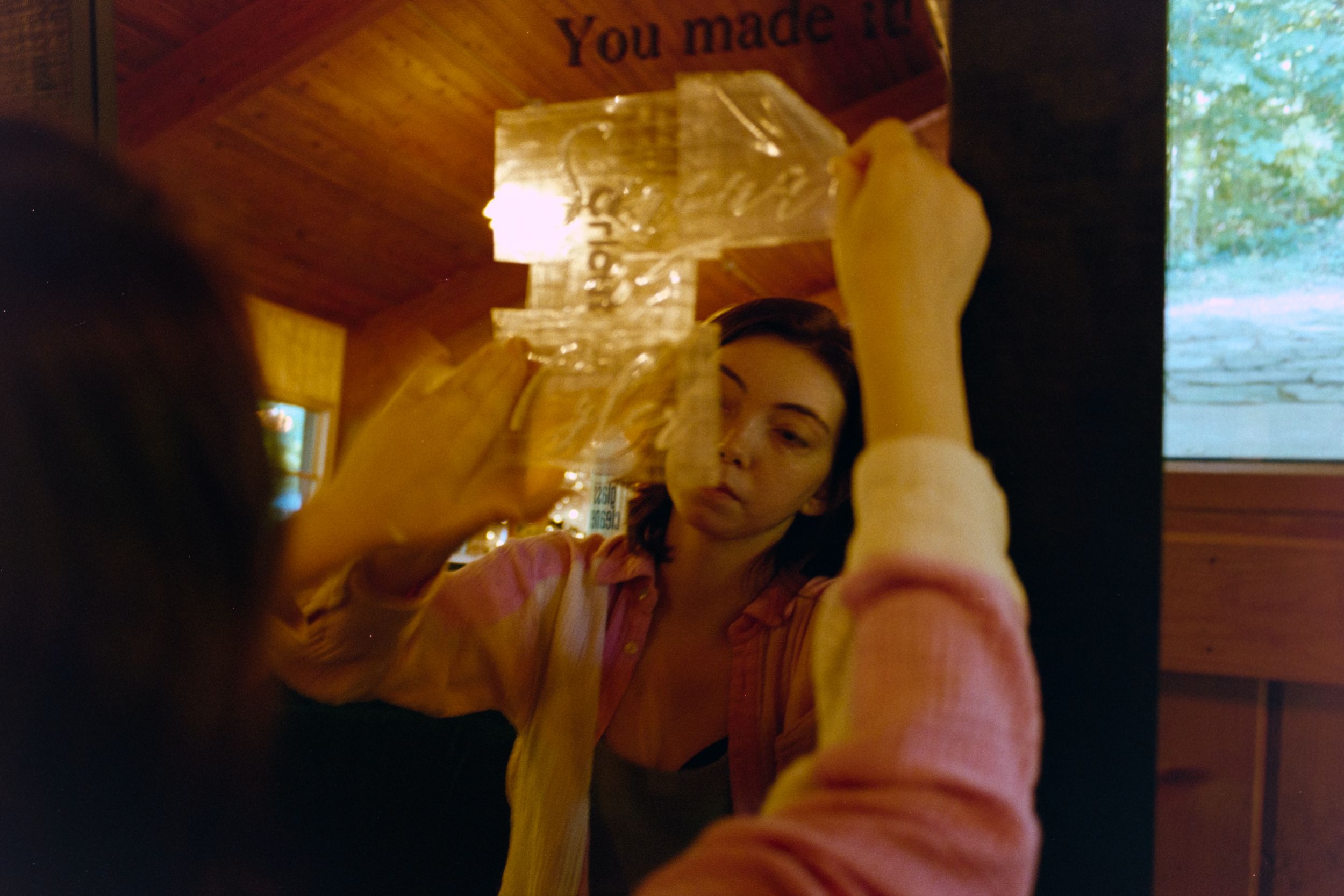 Woman in pink shirt adjusts plastic wrap on mirror with motivational text reflection.