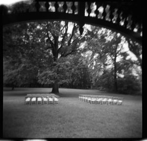 Black and white Holga photo of rows of white chairs set up outdoors under large trees for a ceremony.