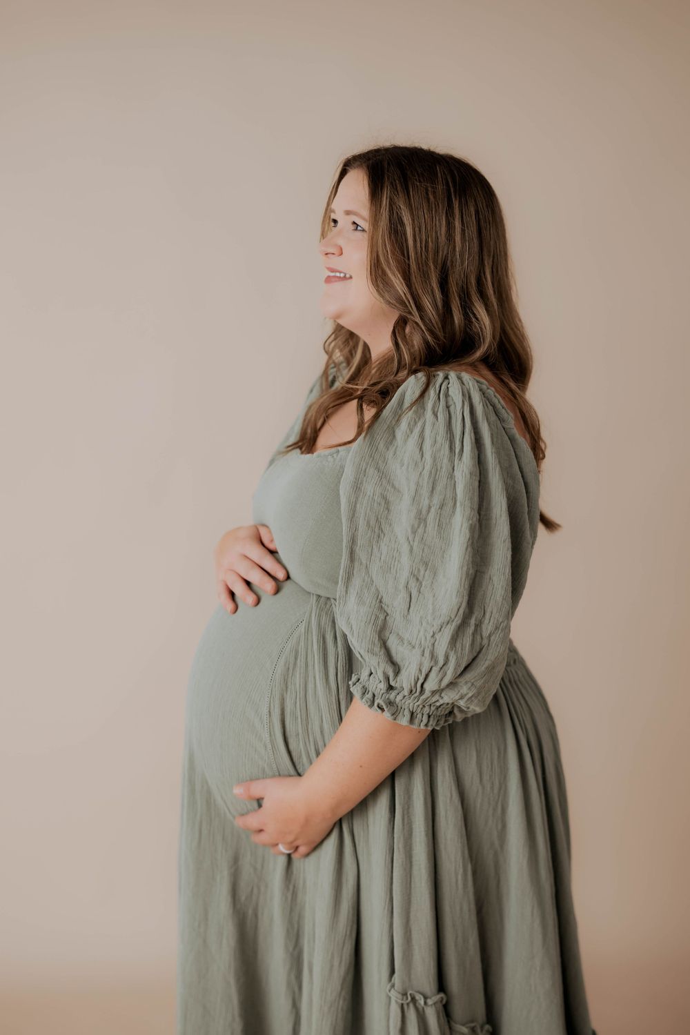 Side profile of someone in a sage green maternity dress with puff sleeves posing against beige background.