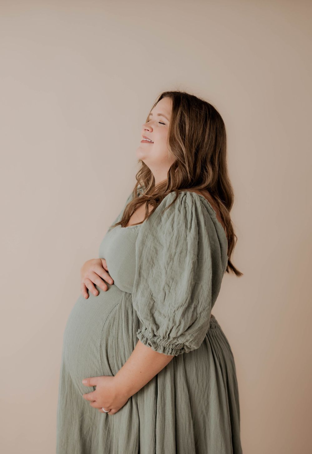 Side view of an expectant mother in a sage green dress smiling with hands on her pregnant belly against beige background.