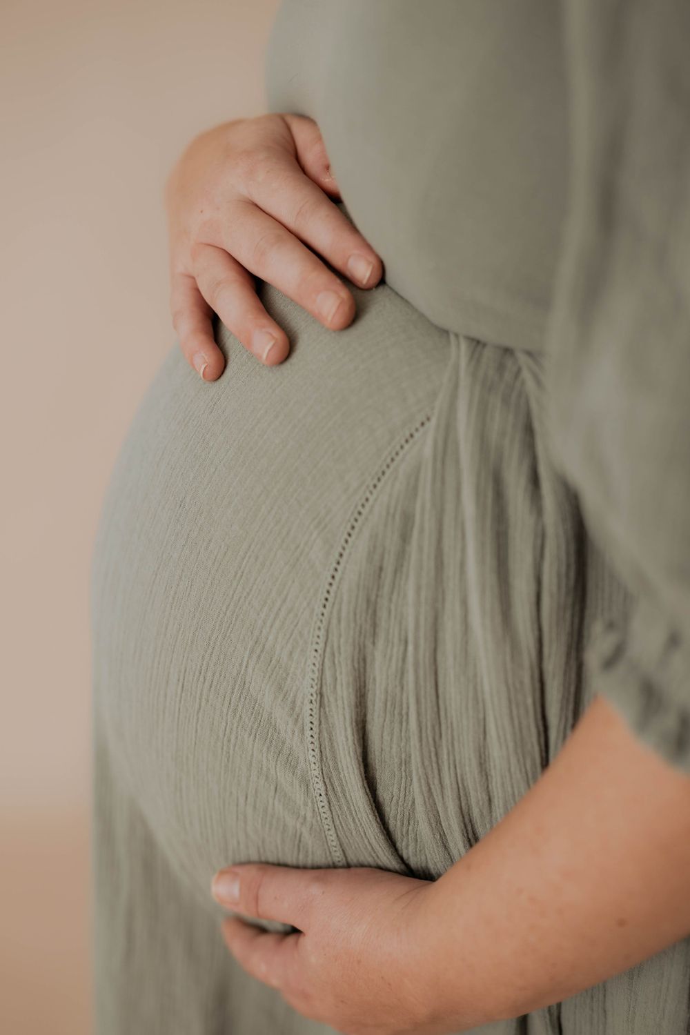 Close up view of a pregnant belly in a sage green crinkle fabric dress with soft gentle hands cradling the bump.