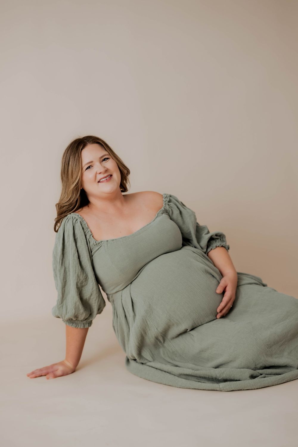 A maternity photo featuring someone in a sage green dress with puff sleeves posing against a neutral backdrop.