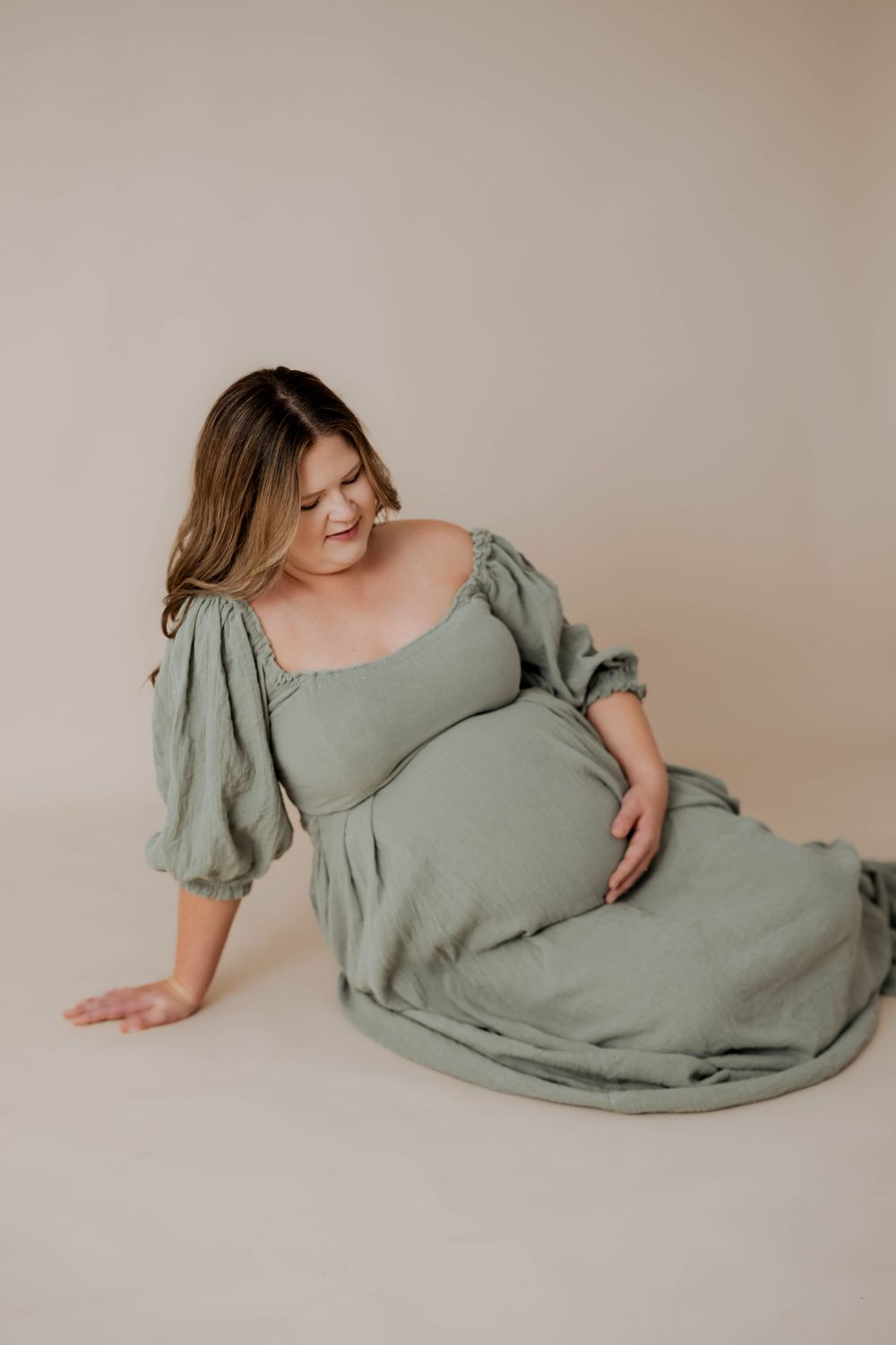 A maternity portrait features a pregnant woman in a sage green dress with puff sleeves sitting elegantly on a beige background.