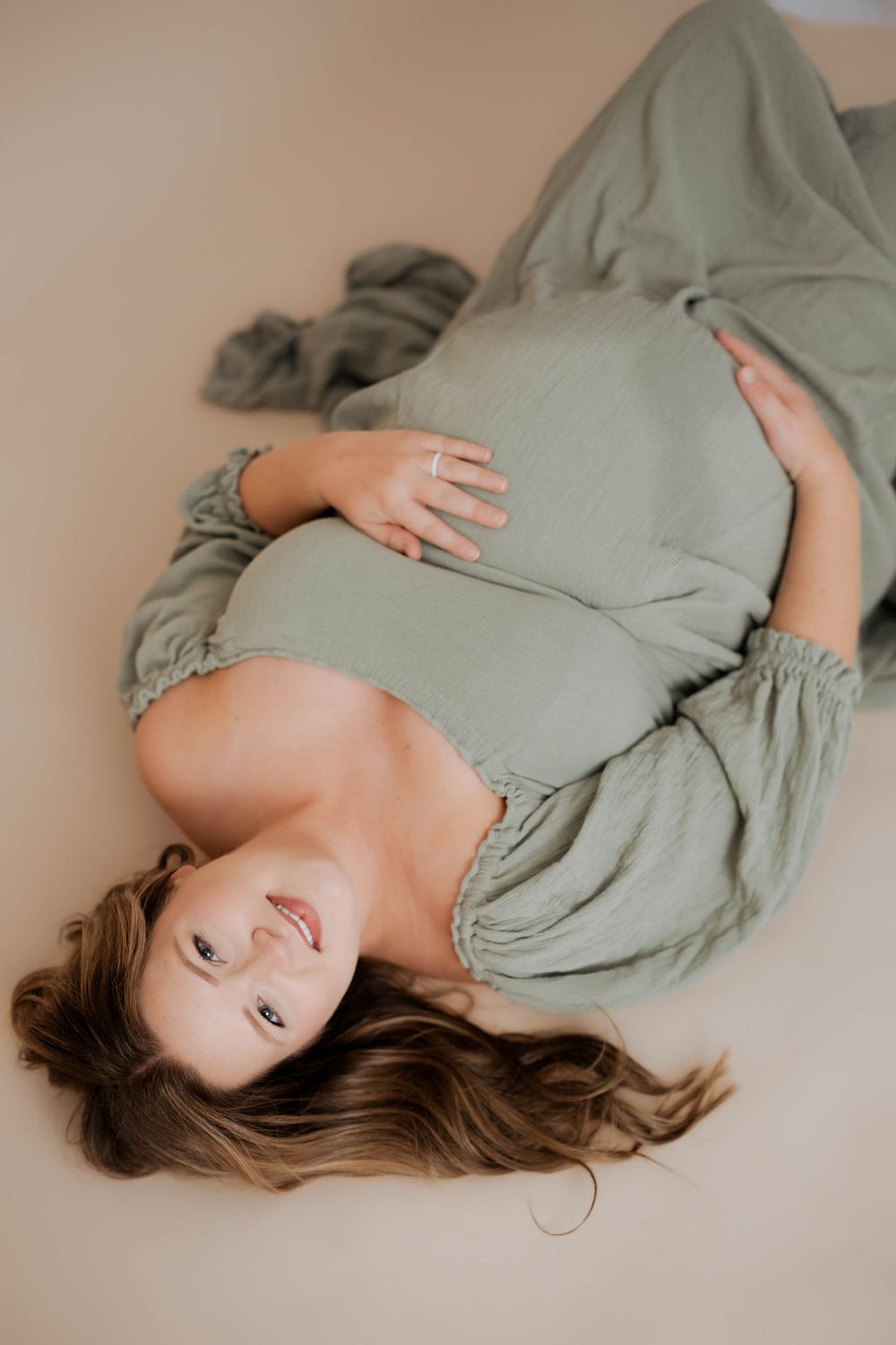 A maternity photo shows a pregnant woman lying down in a sage green dress against a neutral backdrop.