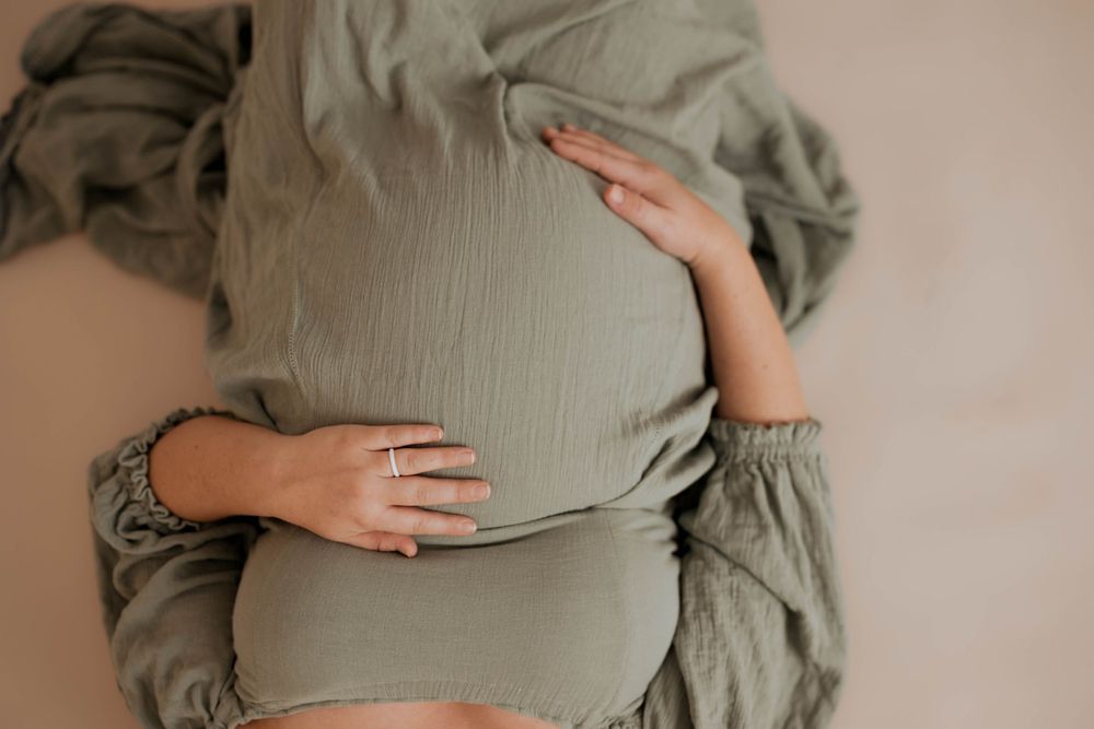 Hands gently cradle a pregnant belly wearing a sage green dress against a soft neutral background.